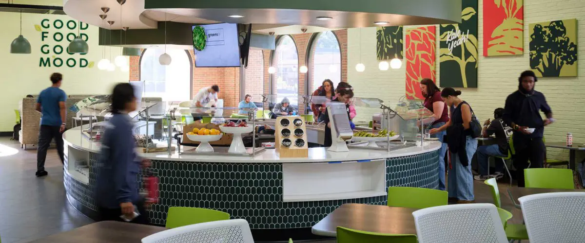 people serving themselves at a circular salad bar labeled "greens," surrounded by tables and chairs with colorful, abstract wall art in the background.