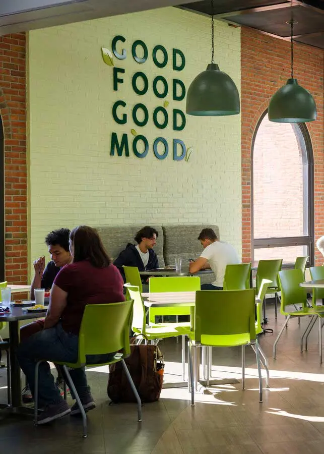 green chairs and hanging pendant lights, where people are seated and engaged in conversation or working on laptops, under a wall sign that reads "GOOD FOOD GOOD MOOD."