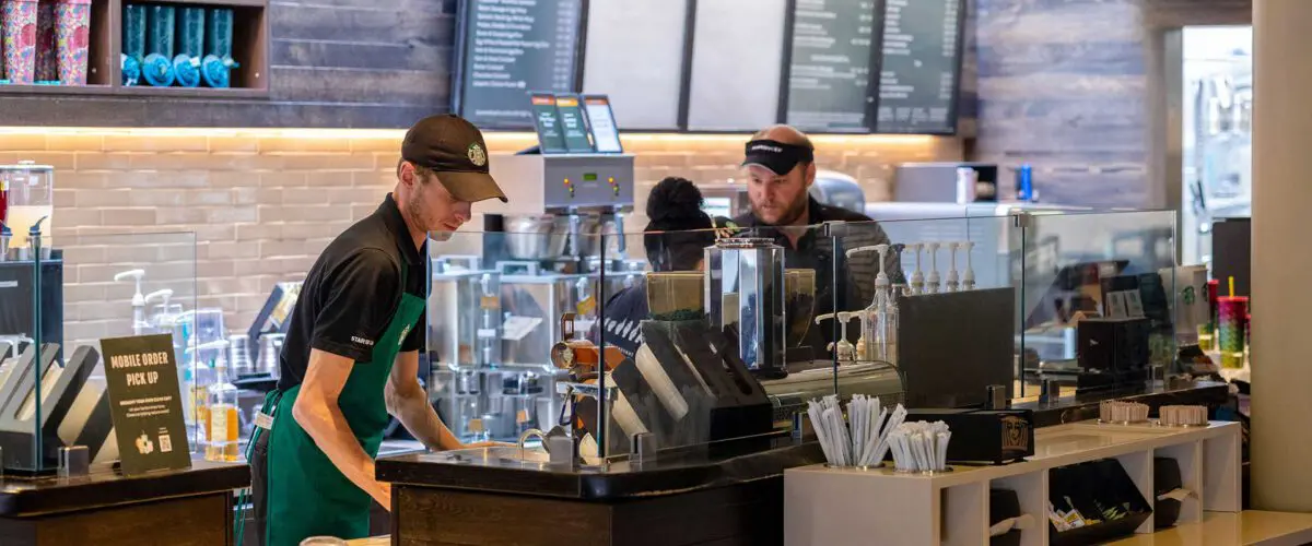 two baristas behind the counter preparing drinks in a campus coffee shop with menu boards and various coffee equipment.