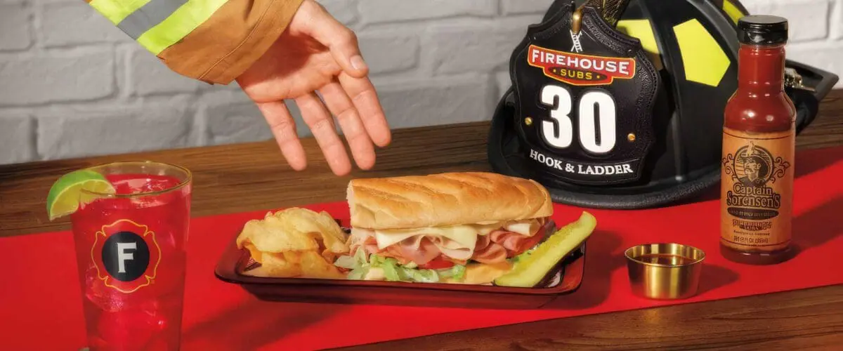 Firehouse Subs sandwich with chips and pickle on a tray beside a firefighter helmet and hot sauce bottle.