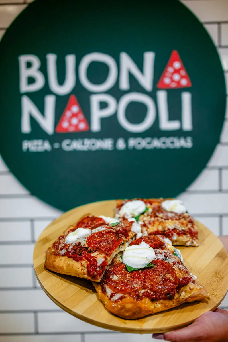 Hands holding pizza on board with Buona Napoli sign in background