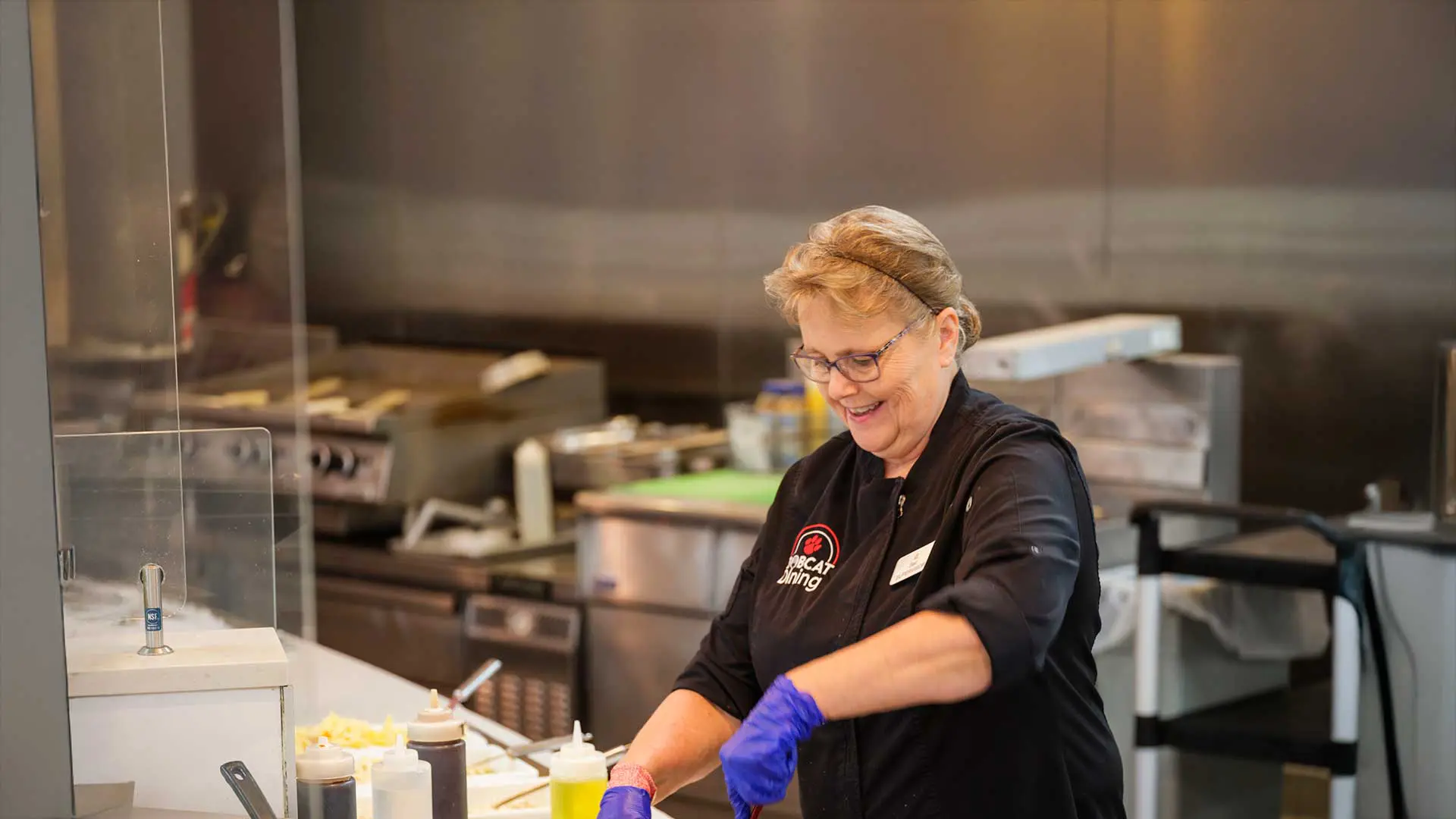a smiling female chef wearing blue gloves and a black uniform, preparing food in two pans