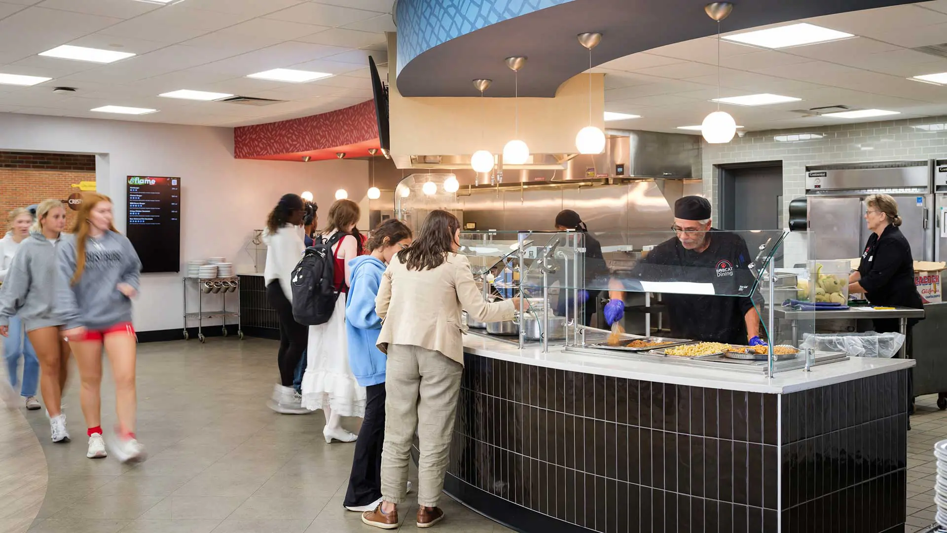 a cafeteria setting where people are waiting in line as a food service worker behind a counter serves meals.