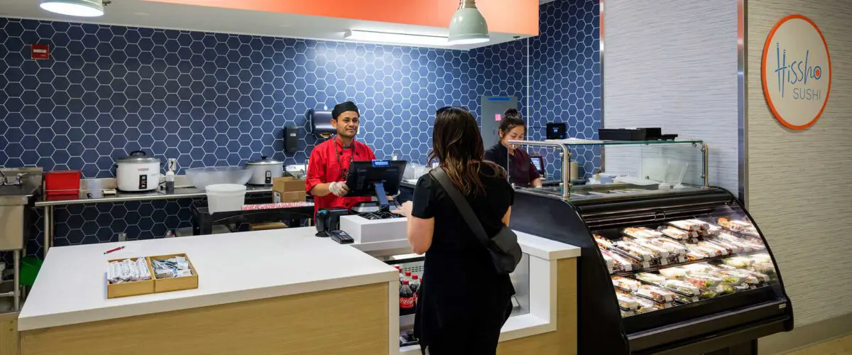 Woman ordering sushi at a modern sushi counter inside a brightly lit restaurant with blue hexagonal wall tiles