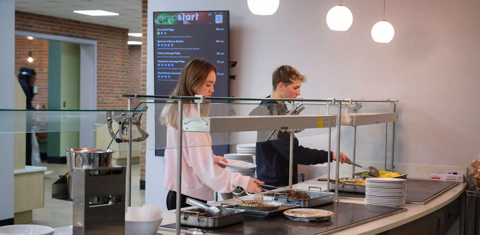 two young people serving themselves food from a cafeteria-style buffet in a modern dining area with bright overhead lighting.