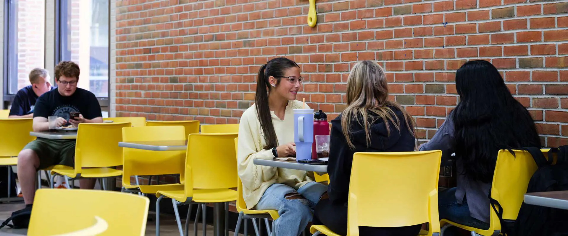 a group of people sitting and talking at tables with yellow chairs in a casual dining area with brick walls