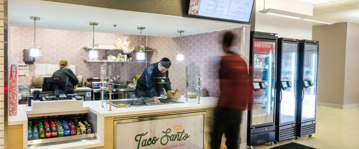 A small, modern food counter named "Taco Santo" inside a building, where two workers prepare food behind the counter while a customer stands in front, likely placing an order.