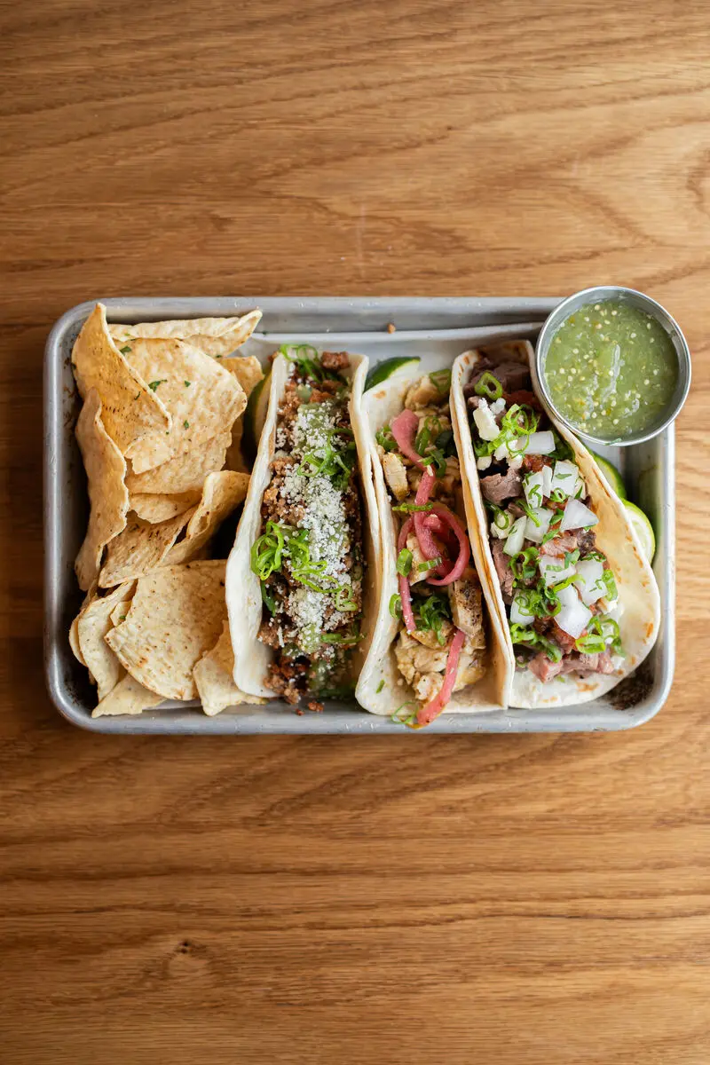 a metal tray with three different tacos filled with various meats and toppings, accompanied by tortilla chips and a small cup of green salsa on a wooden table.