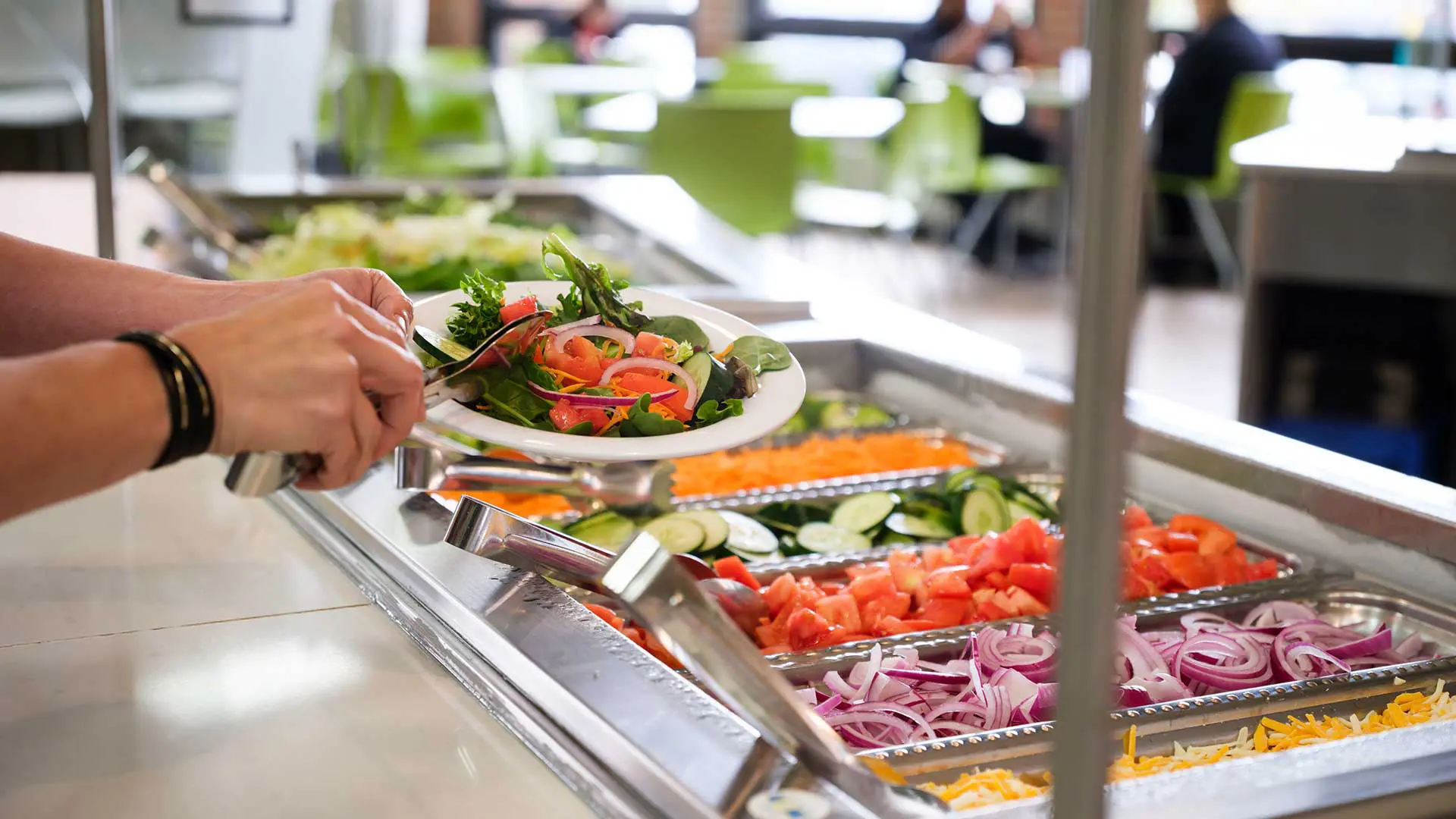 A person is serving fresh vegetables onto a plate from a salad bar in a bright, casual dining area.