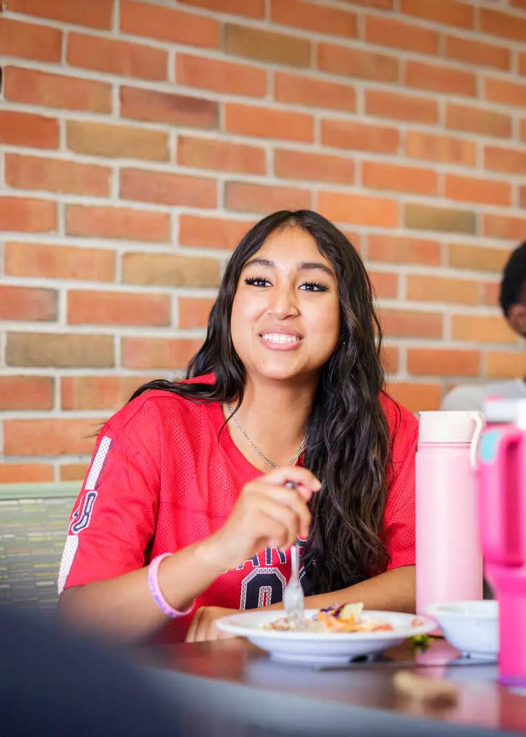 young woman in a red sports jersey sitting at a table indoors with a brick wall background, smiling as she eats a meal with friends.